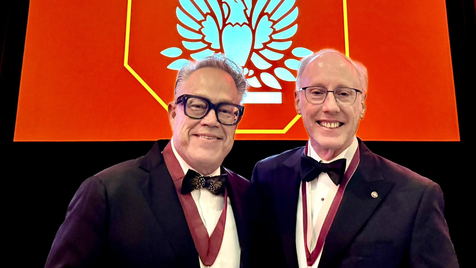 Two men in tuxedos wearing medals stand in front of a red screen displaying an emblem with a column, wings, and a laurel wreath.