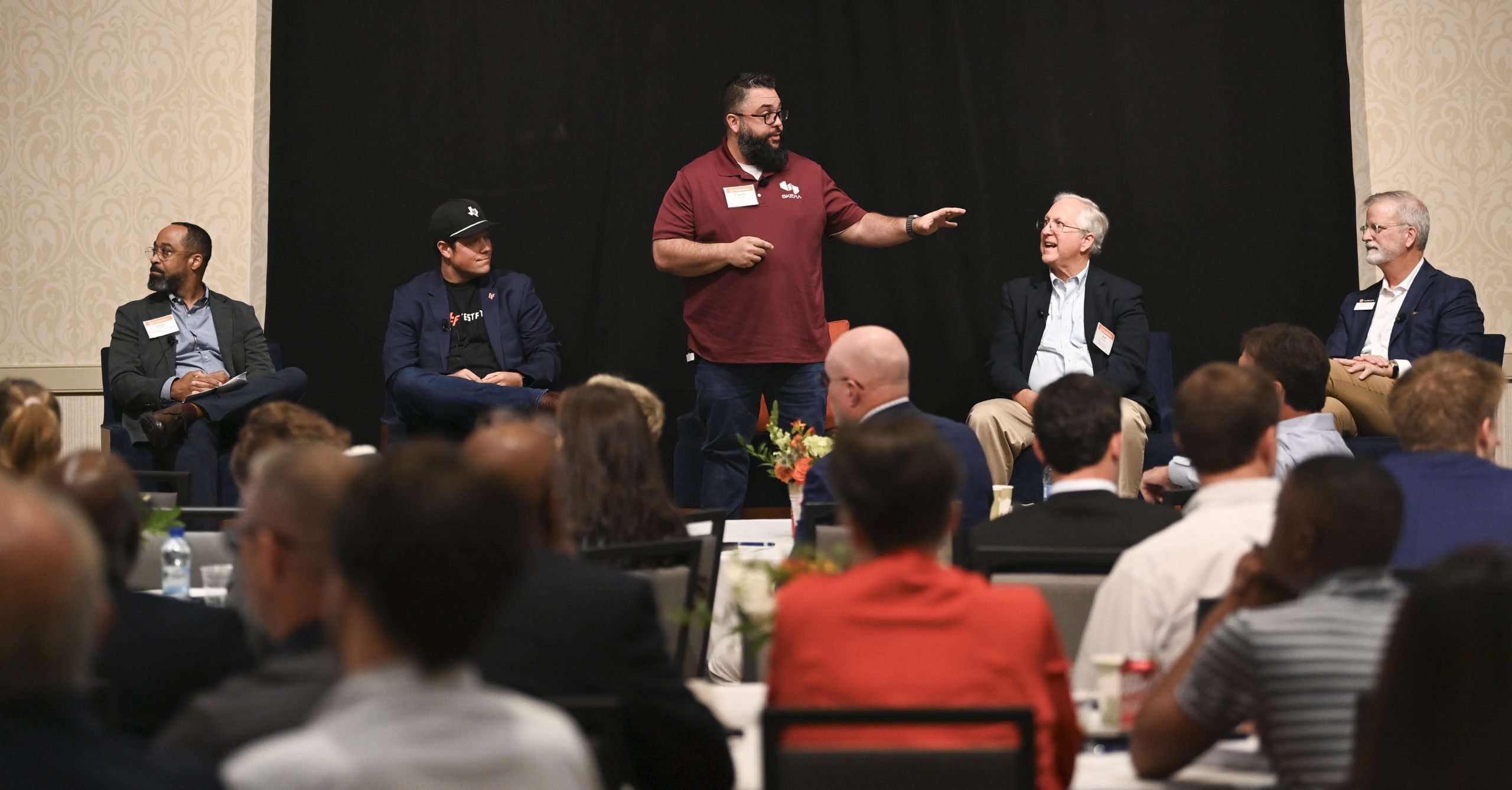 Five men sit on a stage, one standing and speaking, addressing an audience at a formal indoor event or panel discussion.
