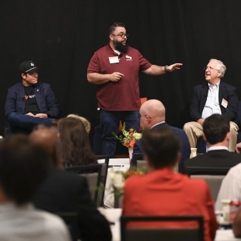 Five men sit on a stage, one standing and speaking, addressing an audience at a formal indoor event or panel discussion.