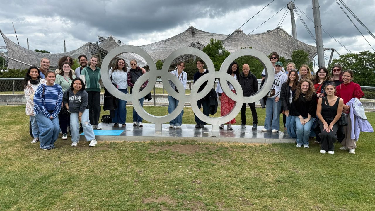 A group of people pose outdoors in front of large Olympic rings, with modern tent-like structures and cloudy skies in the background.