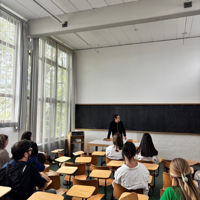 A teacher stands at a desk in front of a chalkboard, speaking to students seated in rows of wooden desks in a classroom with large windows.