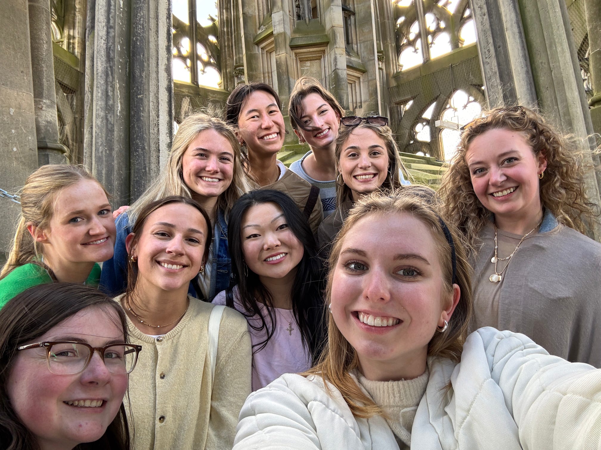 A group of ten young women smile for a selfie in front of an ornate, gothic-style stone structure.