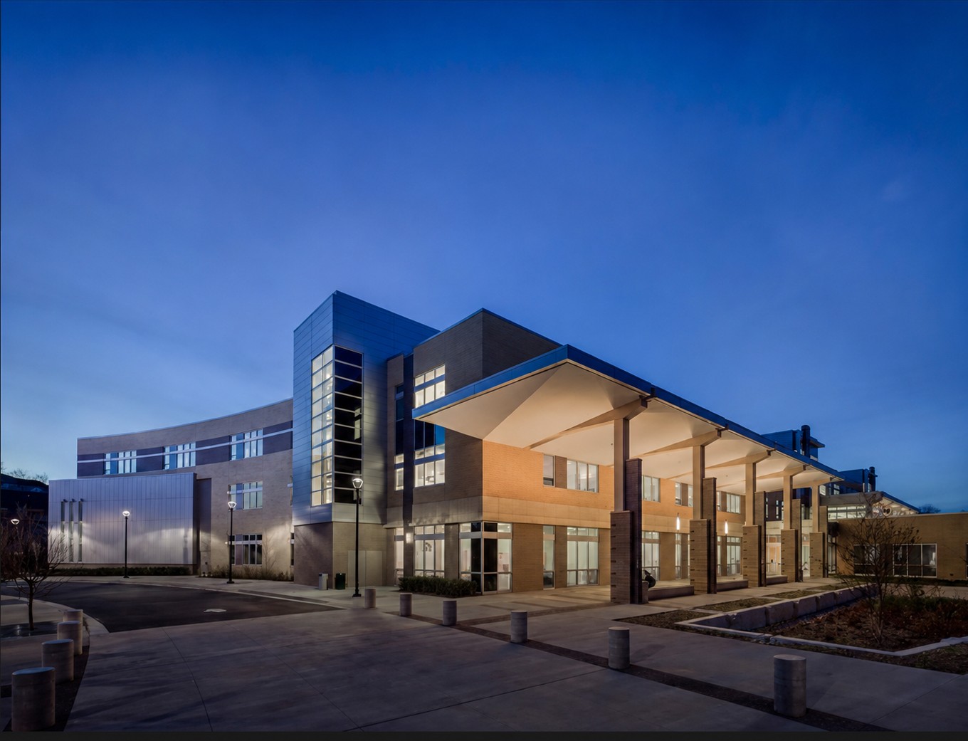 Modern school building with large windows and a covered entrance, illuminated at dusk under a clear blue sky.