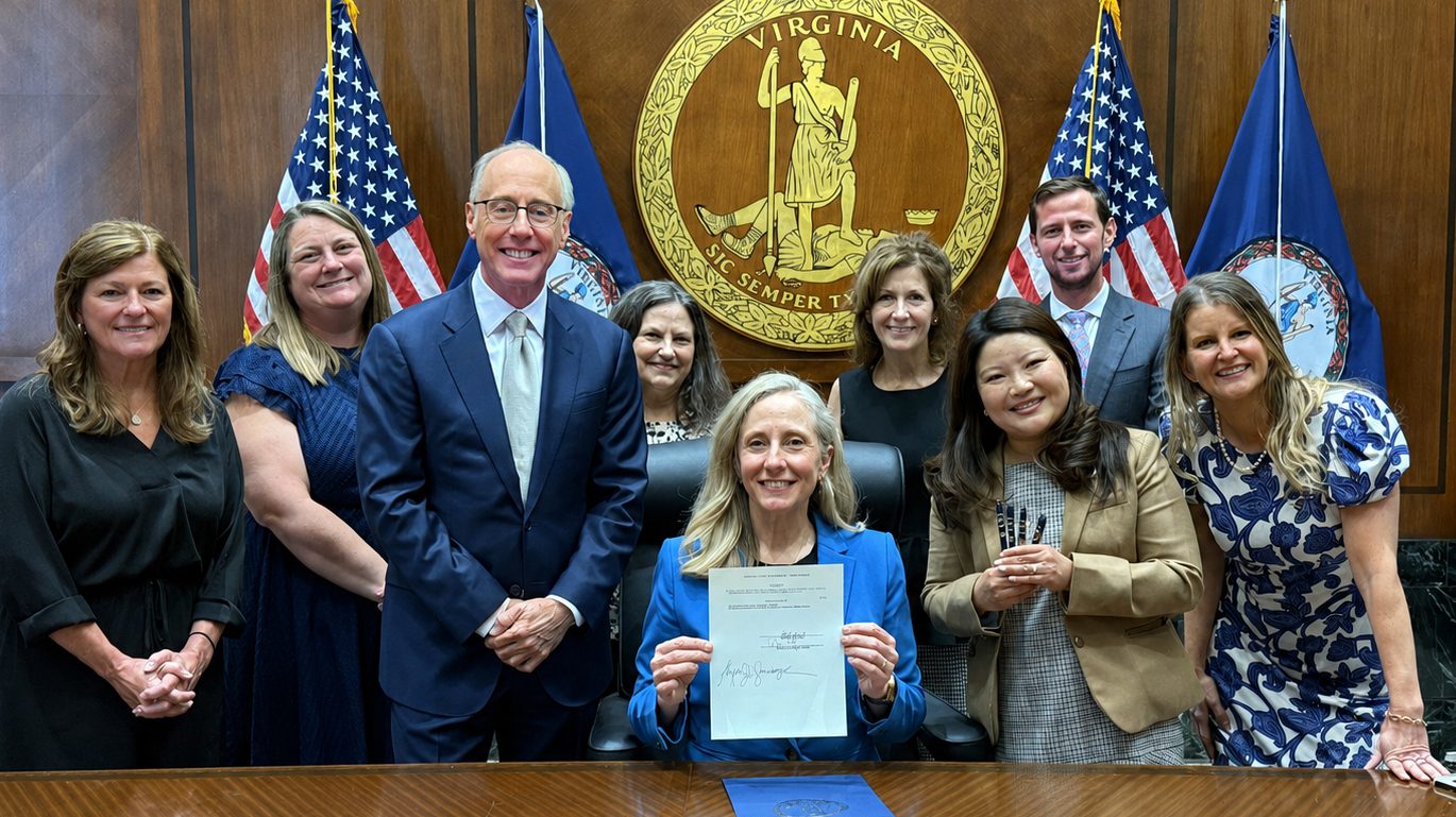 A group of nine people pose around a desk; one person seated at the center holds up a signed document. The Virginia state seal and flags are displayed in the background.