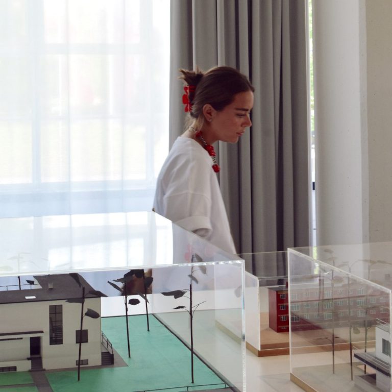 A person stands near a window with gray curtains, beside architectural models displayed on tables in a brightly lit room.
