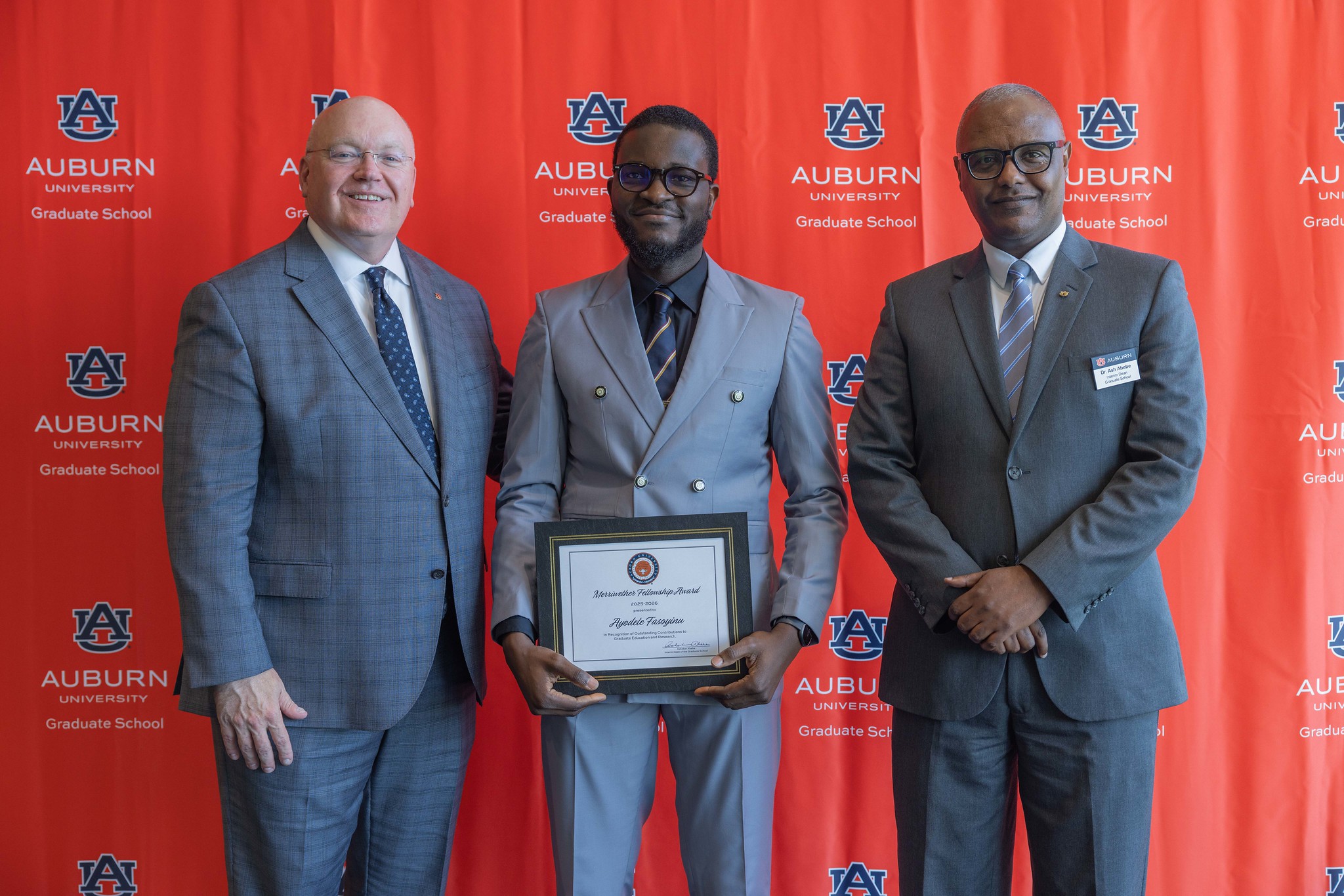 Three men in suits stand in front of an Auburn University Graduate School backdrop; the man in the center holds an award certificate.