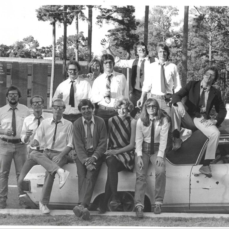 A group of thirteen young adults in formal and semi-formal attire pose together on and around a car parked outside, with trees and a brick building in the background.
