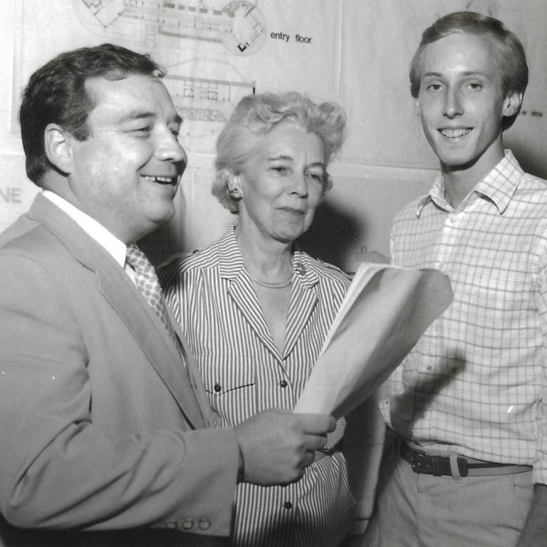 Three people stand together indoors; one man in a suit holds papers and smiles, while a woman and a younger man in casual attire look toward the camera. Blueprints are visible in the background.