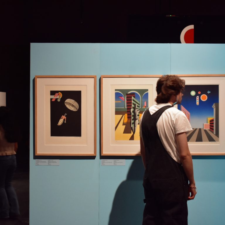 A person in overalls stands in front of framed abstract artworks on a light blue wall in a gallery, while others view art in the background.