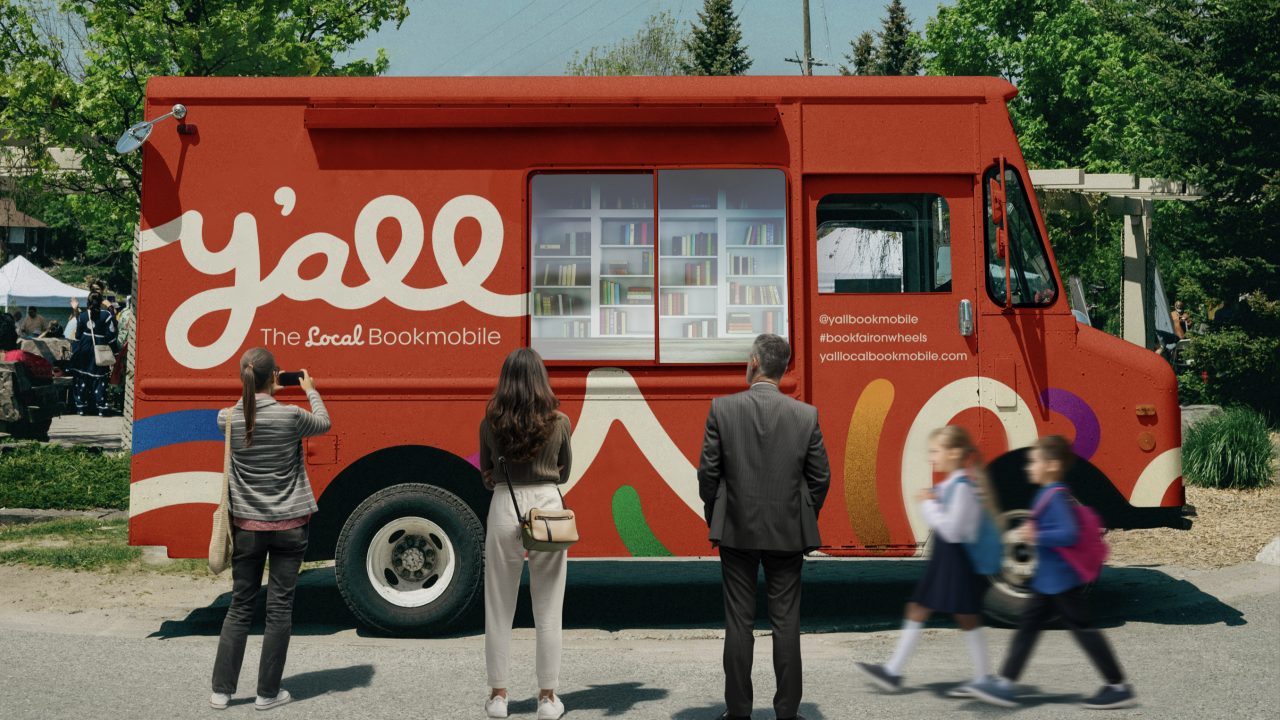 A red bookmobile labeled yall The Local Bookmobile is parked outdoors. Three adults stand facing it, while two children walk past in the foreground.