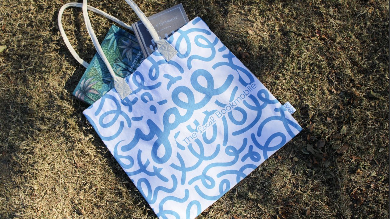 A blue and white tote bag with abstract designs and “The Local Bookmobile” text sits on grass, partially filled with books.