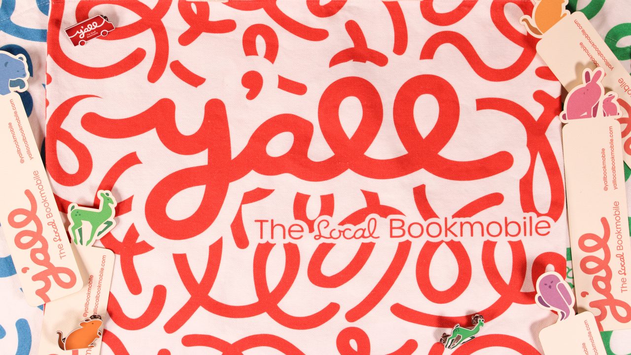 A white cloth with “y’all” and “The Local Bookmobile” printed in red, surrounded by bookmarks and small animal cutouts.