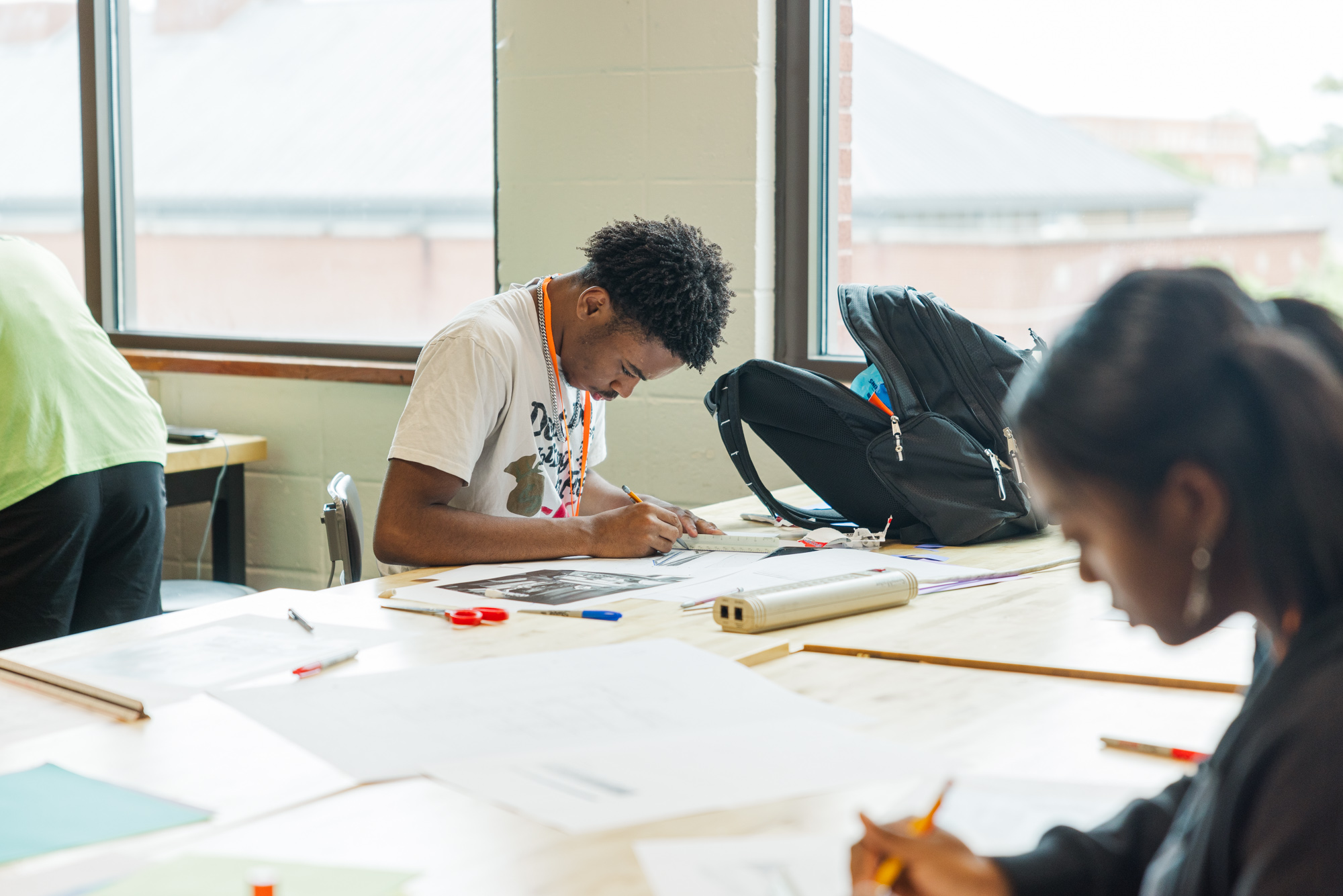 A student sits at a table in a classroom, intently working on a drawing, with art supplies and papers spread out around him.