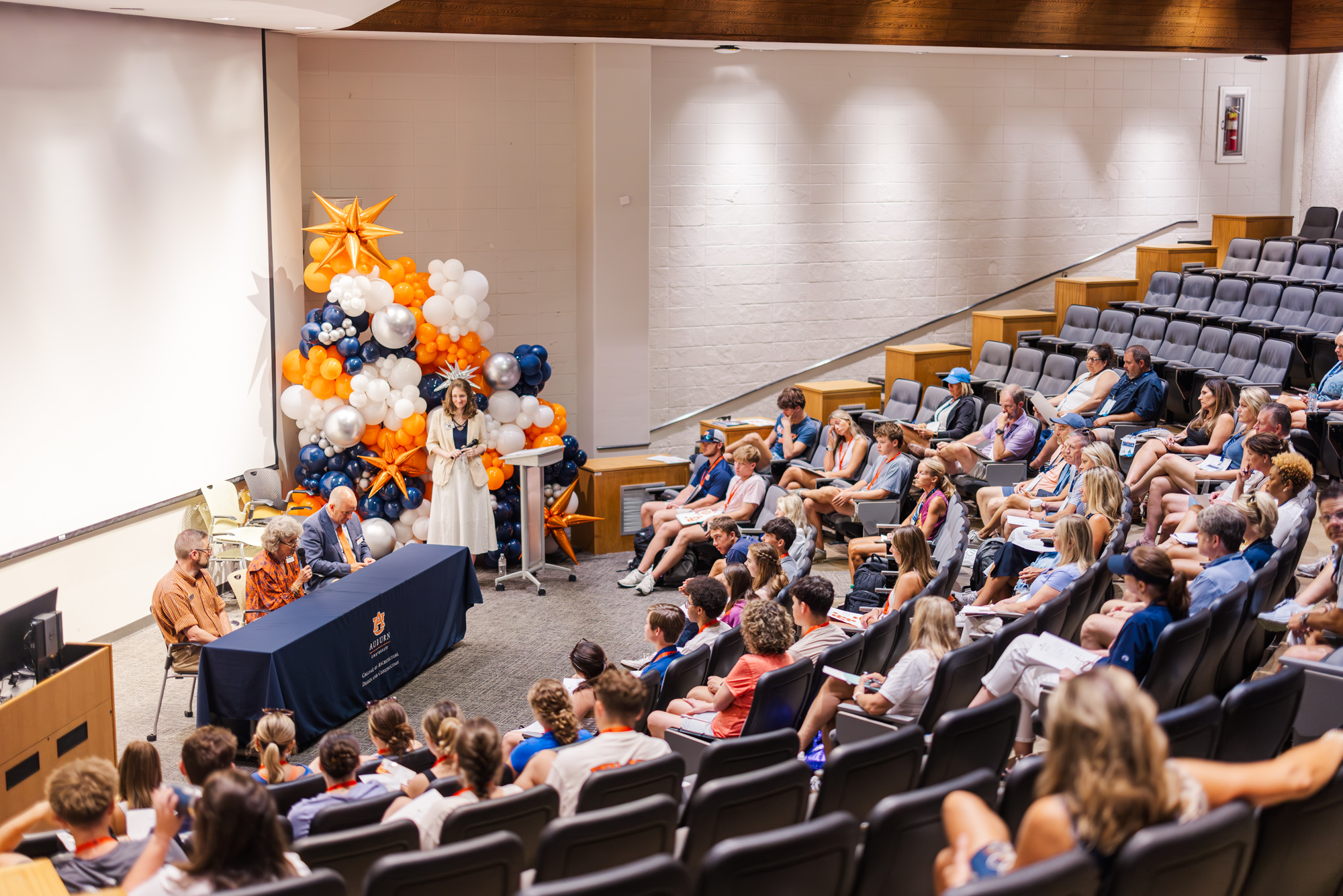 A panel of five people sits at a table in front of an audience in a lecture hall, with balloon decorations behind them.