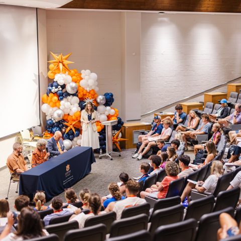 A panel of five people sits at a table in front of an audience in a lecture hall, with balloon decorations behind them.