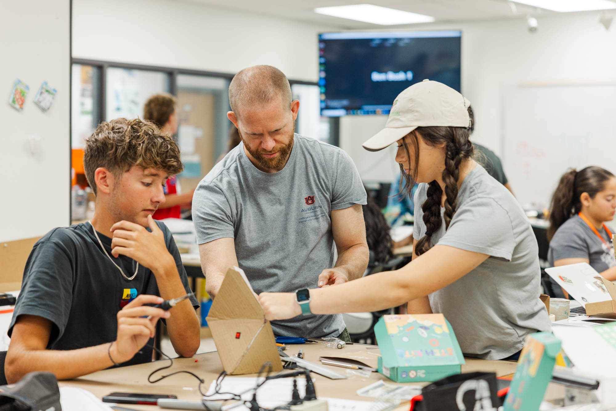 A teacher assists two students with an electronics project at a table in a classroom, while other students work in the background.