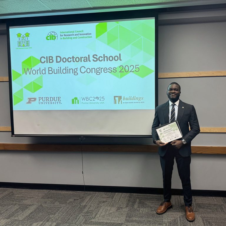 A man in a suit holds a certificate and stands in front of a presentation screen displaying CIB Doctoral School World Building Congress 2025 at Purdue University.