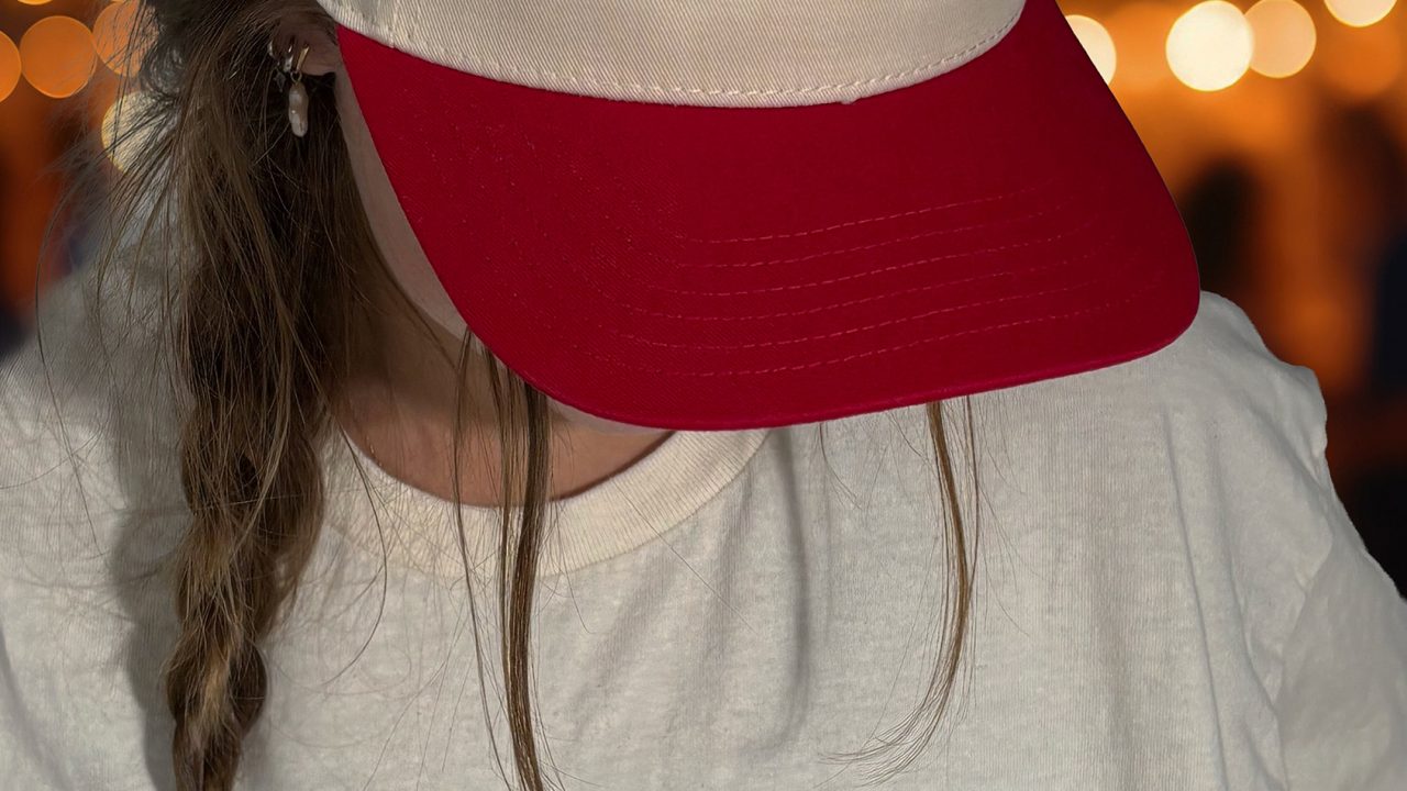 Person wearing a cream and red Club 7 hat and matching Club 7 Live + Dive t-shirt against a blurred background with string lights.