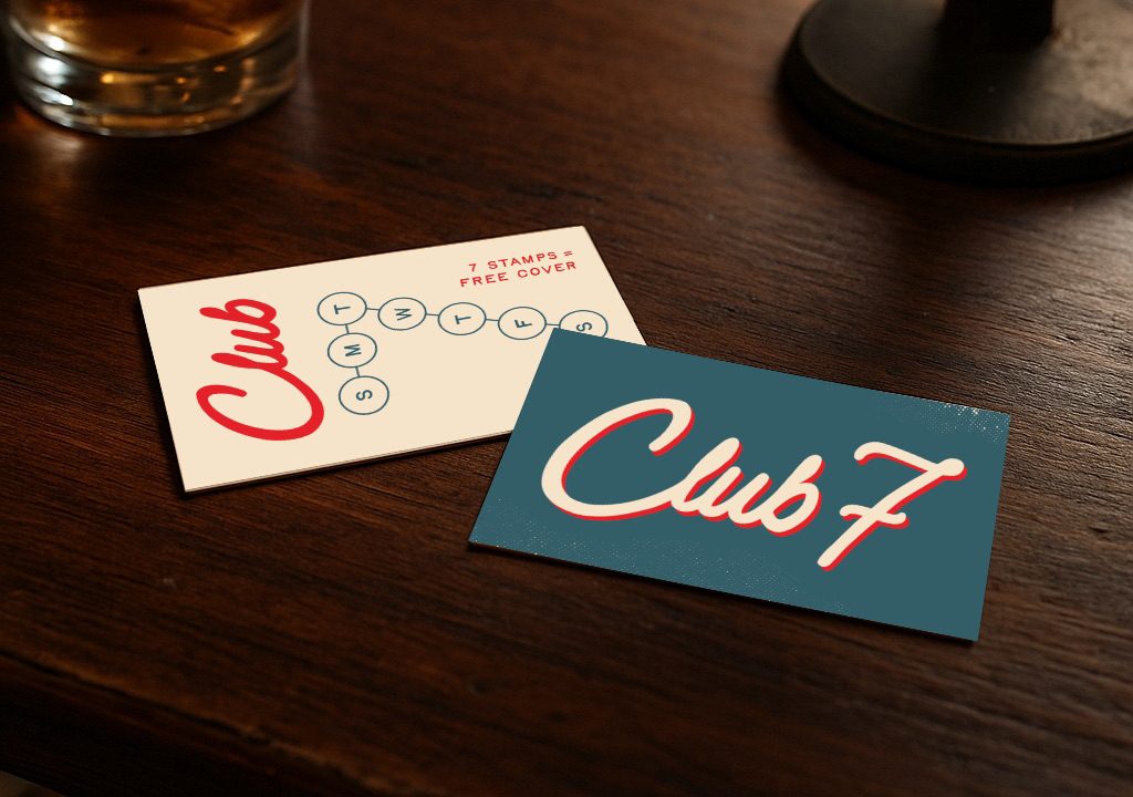 Two Club 7 cards, one a loyalty punch card and the other a logo card, are placed on a wooden table next to a glass of brown liquid with ice.