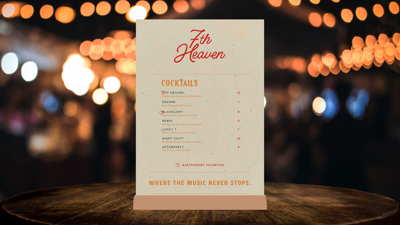 A cocktail menu titled 7th Heaven displayed on a wooden table, listing various drinks with prices, set against a background of blurred warm lights.