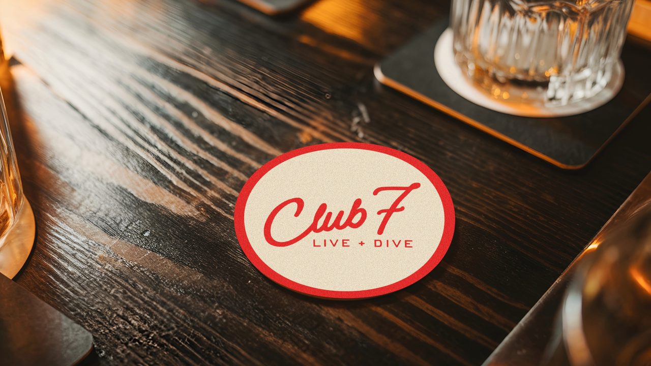 A round coaster with Club 7 LIVE + DIVE in red text sits on a dark wooden table next to glasses.