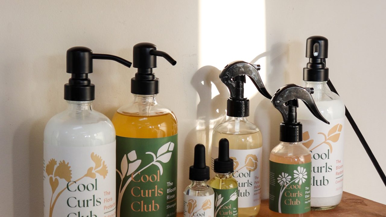 A set of Cool Curls Club hair care products arranged on a wooden shelf below a checkered black-and-white wall.