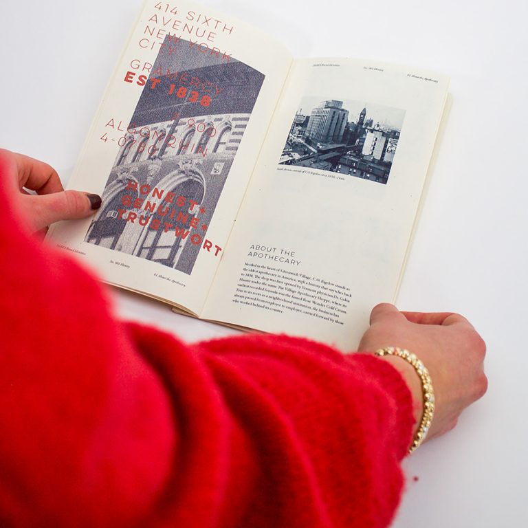 A person in a red sweater is holding open a book displaying a graphic page on the left and a black-and-white city photo with text on the right.