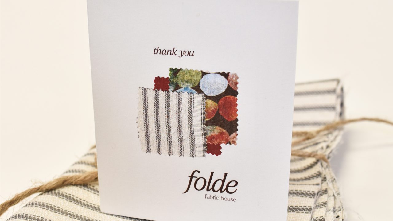 A thank you card with folde fabric house logo and fabric swatches stands on top of folded striped fabric with a string.