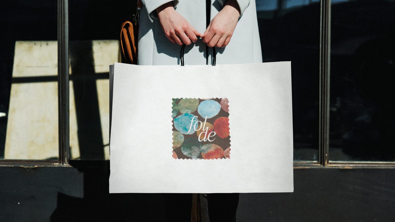 A person holds a large white shopping bag with an abstract colorful design and the word fol de printed on the front.