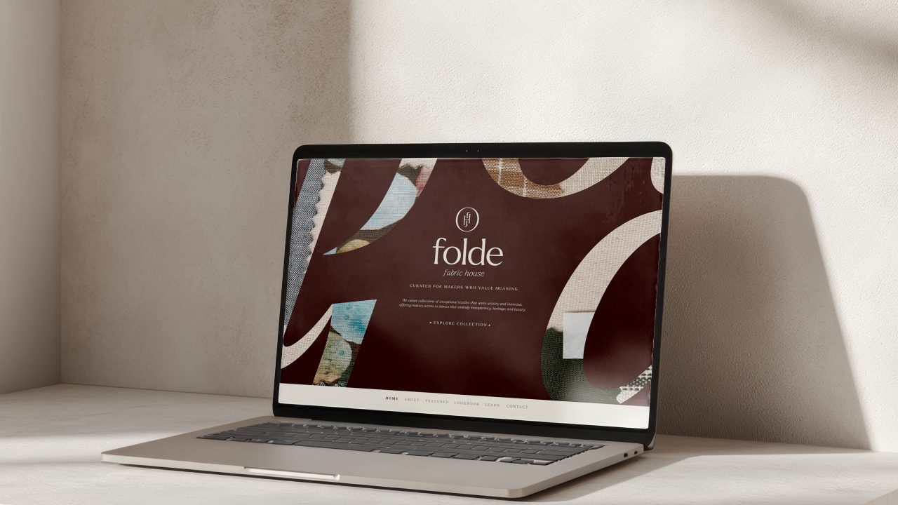 A laptop on a light-colored stone surface displays a website with the text folde, abstract designs, and neutral tones, against a sunlit, textured wall.
