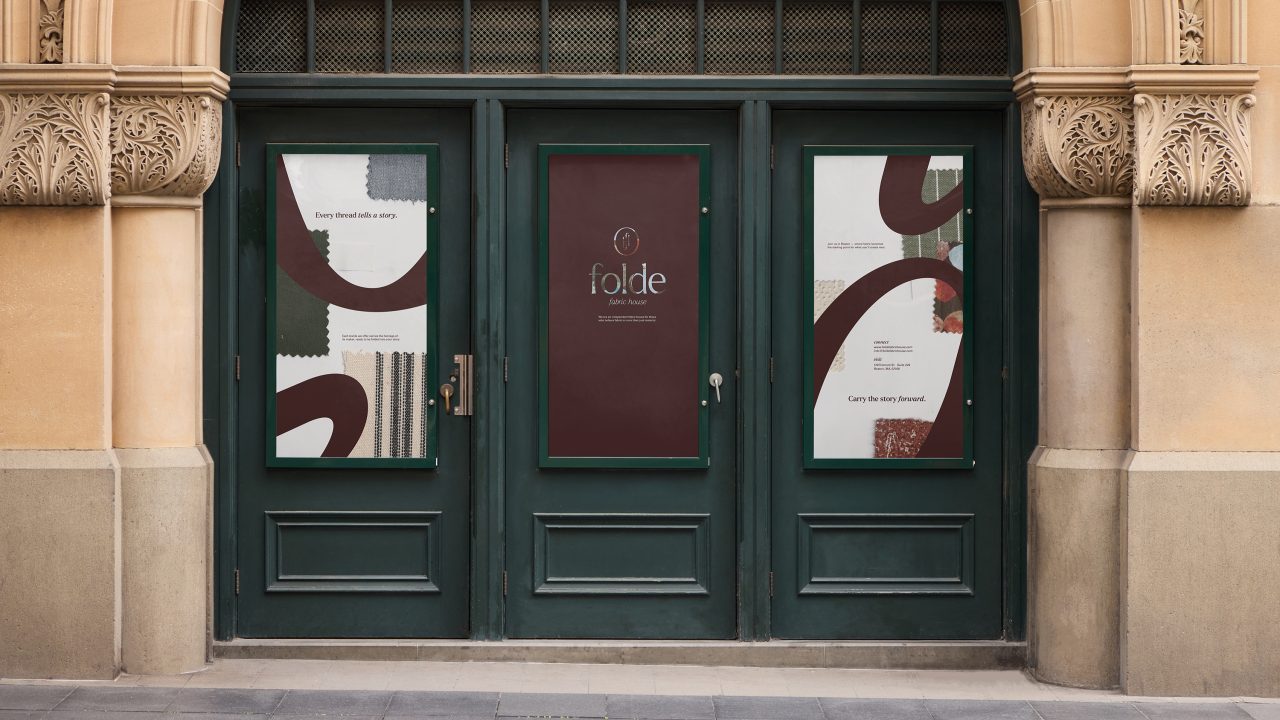 Storefront with three green-framed doors; center door displays folde branding, side doors feature abstract designs and fabric swatches with brief promotional text.