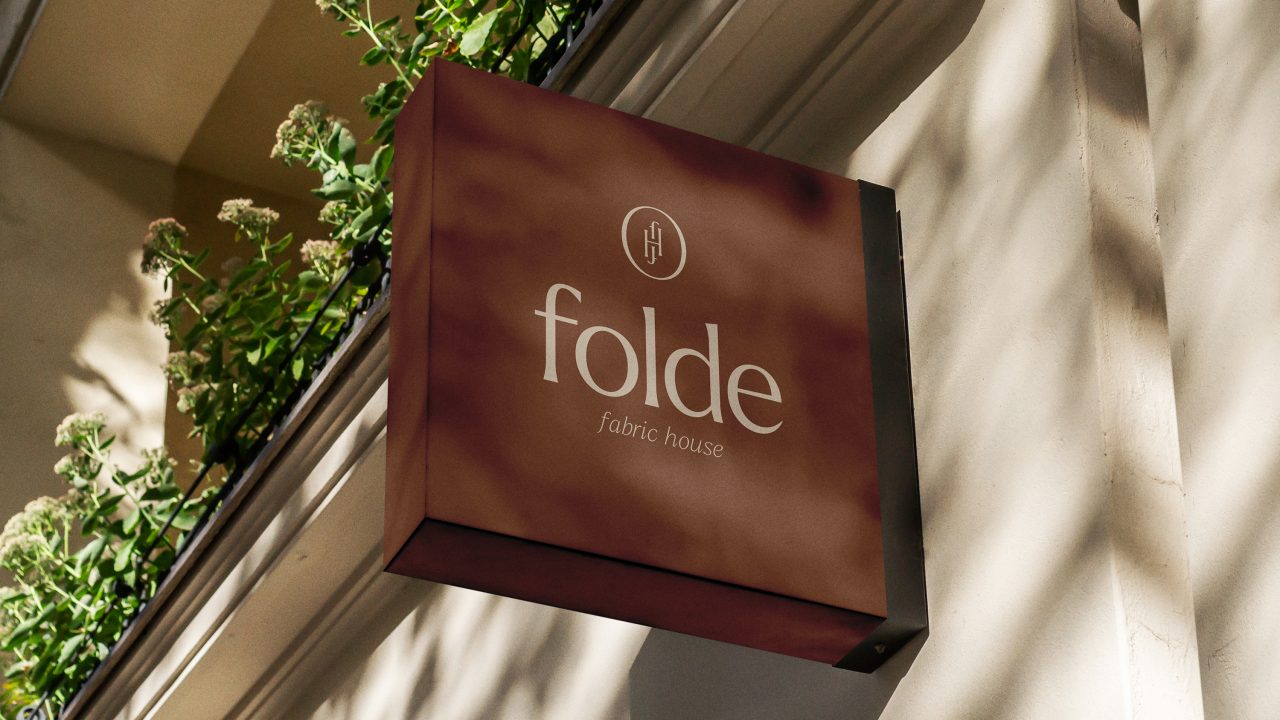A brown fabric house sign reads folde fabric house with a circular logo above the text, mounted on a building wall next to green plants.