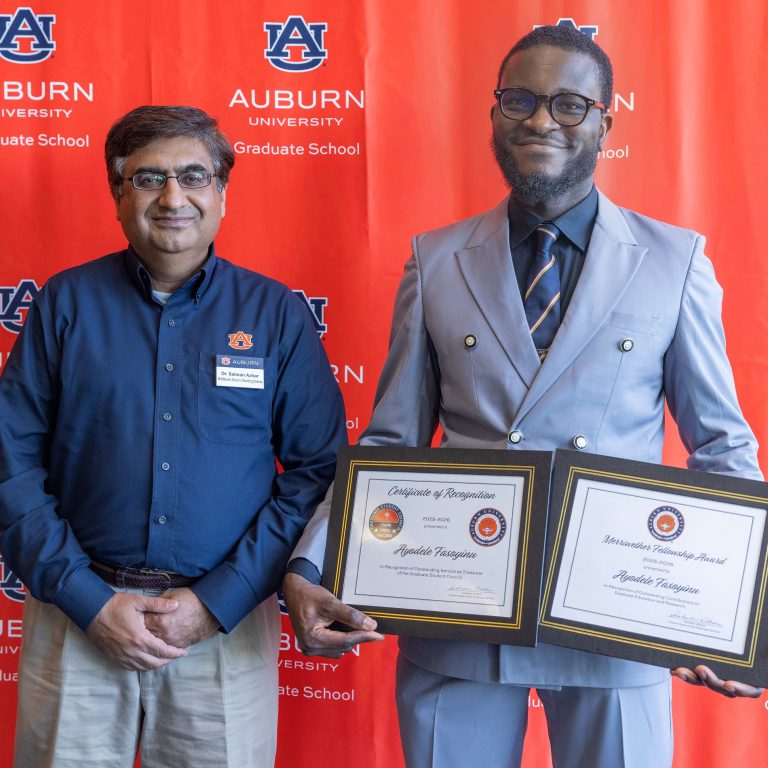 Two men stand in front of an Auburn University Graduate School backdrop; one holds two certificates, while the other stands with hands clasped.