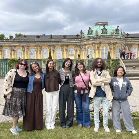 A group of eight people stand in front of a large, ornate yellow building with a green dome under a cloudy sky.