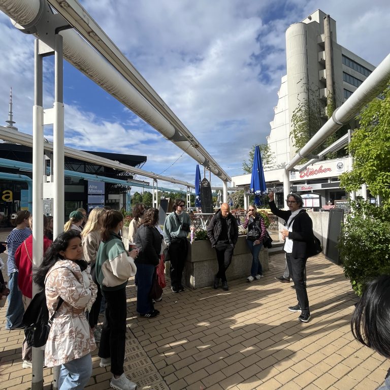 A group of people stands and listens to a person speaking outdoors in a paved urban area with buildings, plants, and a partly cloudy sky.