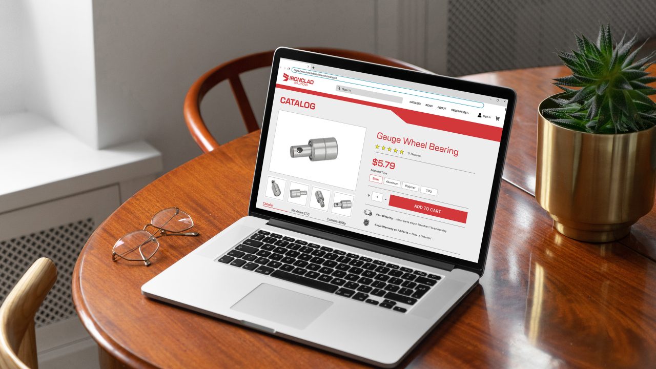 A laptop on a wooden table displays an online store page for a gauge wheel bearing priced at $5.79. Glasses and a potted plant are also on the table.