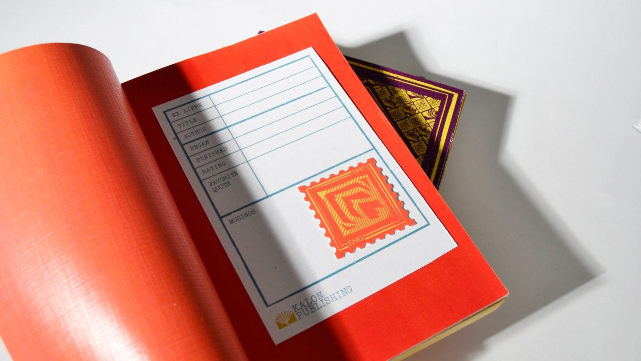 Open book displaying a library card pocket with a red patterned stamp and text fields for title, author, subject, and date. Another closed book with a gold and purple cover is partially visible.