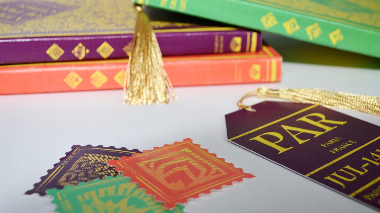 Three decorative books with gold tassels, colorful patterned paper cutouts, and a purple tag labeled PAR are arranged on a white surface.