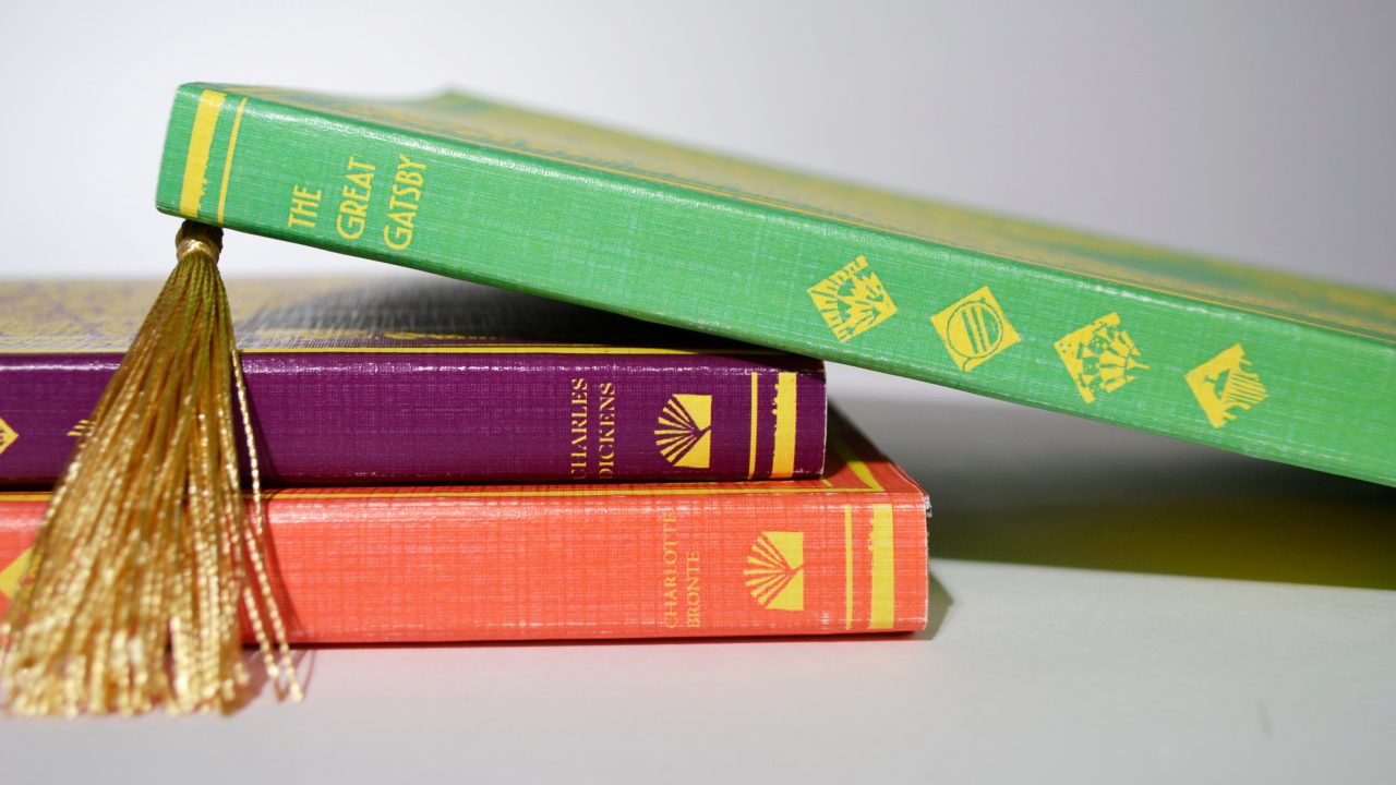 Three hardcover books stacked on a white surface, with The Great Gatsby on top, featuring a gold tassel bookmark.