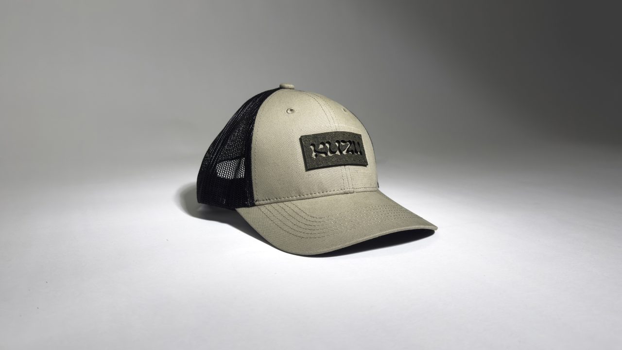 A khaki and black trucker cap with a curved brim and a rectangular patch on the front, displayed on a plain light background.