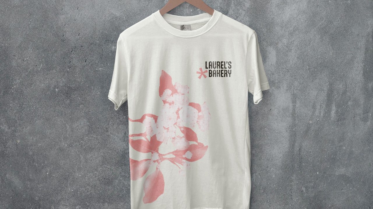 White t-shirt hanging on a wooden hanger against a gray concrete wall, featuring a pink abstract design and the text “Laurie’s Bakery” on the upper left chest.