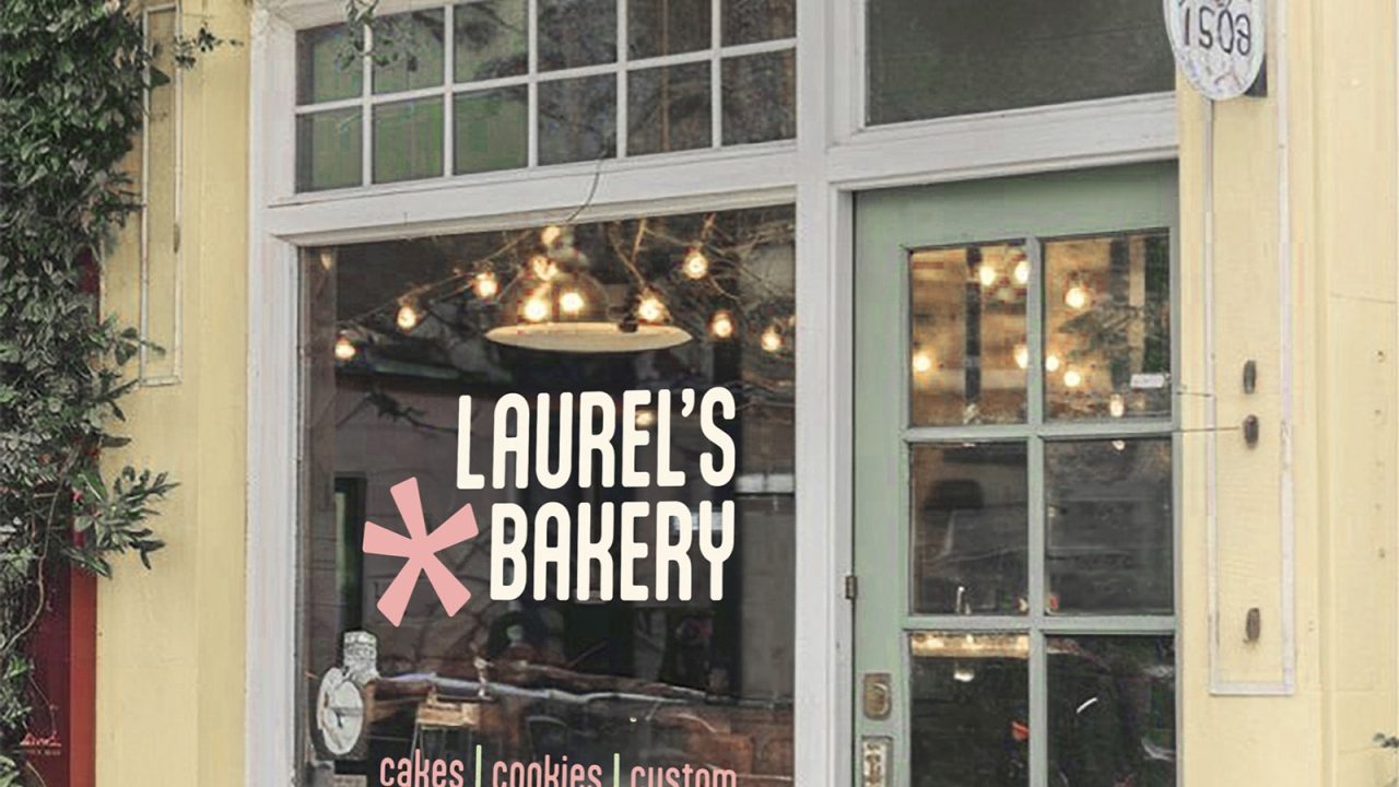 A bakery storefront with ivy-covered yellow walls, a green door, and a window displaying Laurel’s Bakery. Two chairs and a table are placed on the sidewalk outside.