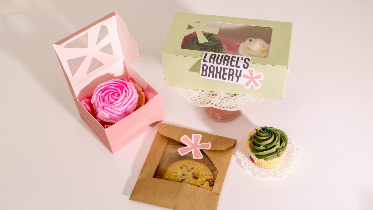 Assorted baked goods from Laurel’s Bakery, including cupcakes in decorative boxes and a cookie in a brown paper bag, displayed on a white surface.
