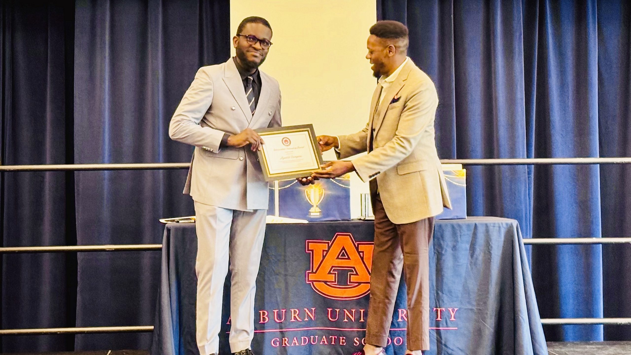 Two men stand on a stage as one presents a framed certificate to the other. Auburn University Graduate School banner is displayed on the table behind them.