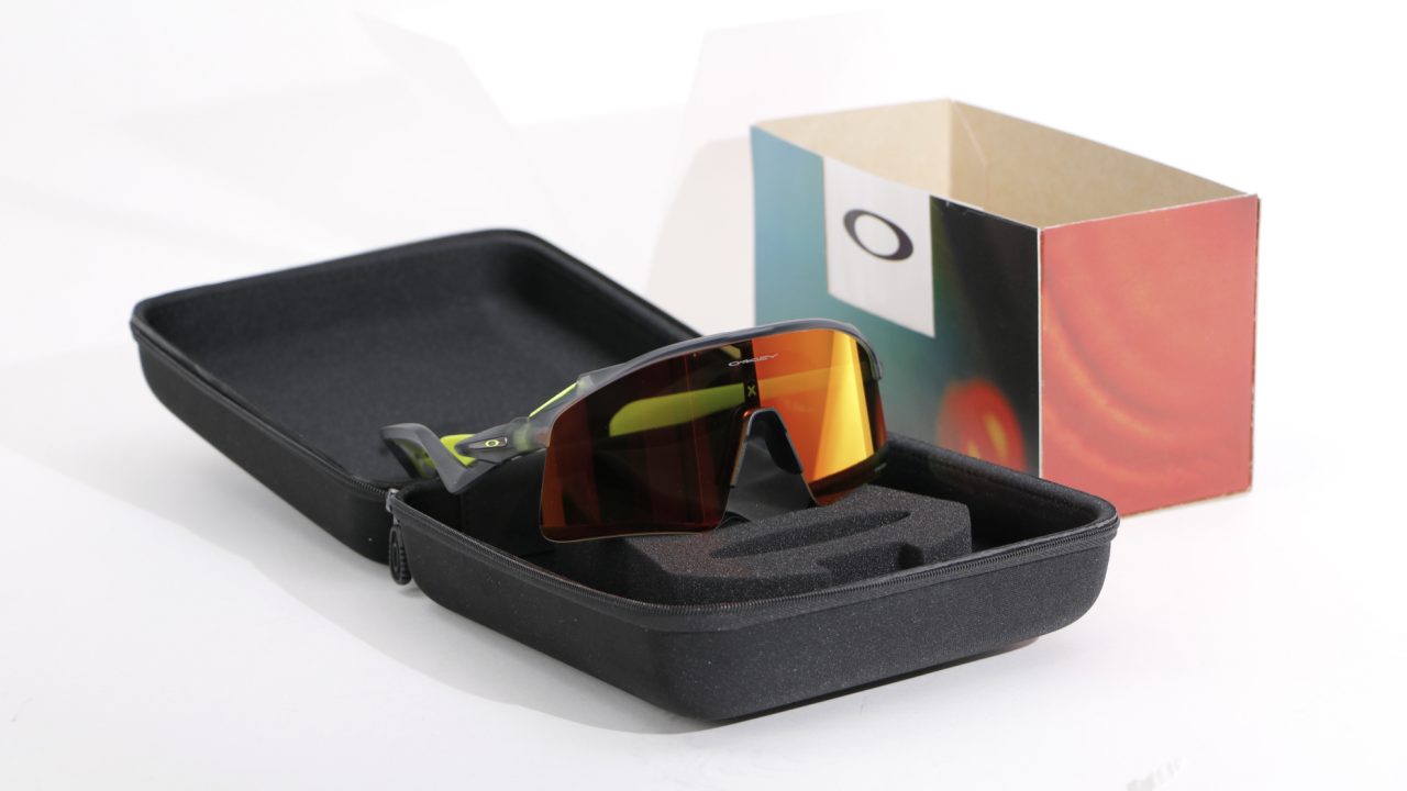 A pair of Oakley sports sunglasses with red-tinted lenses rests in a black case, with an open Oakley box in the background on a white surface.