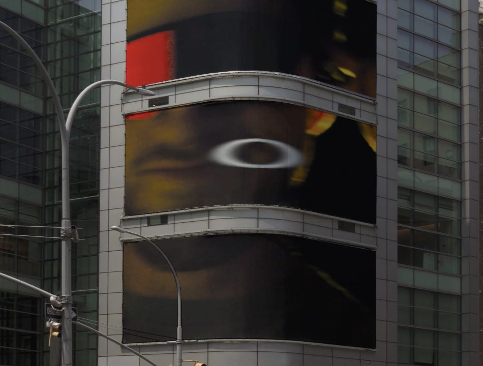A curved digital billboard on a glass building displays an abstract image with dark colors, red, yellow, and white shapes. Cars and pedestrians are visible at the street level below.