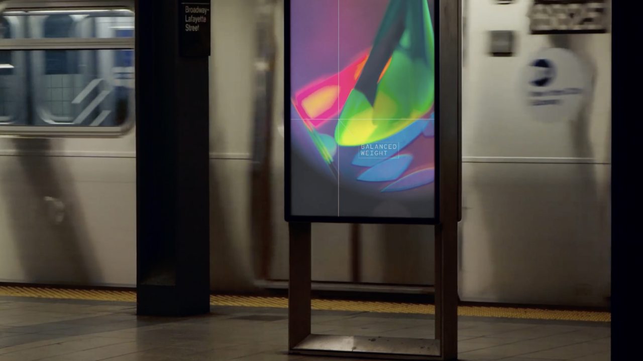 A digital advertisement display with abstract colorful graphics stands on a subway platform as a train passes by in the background.