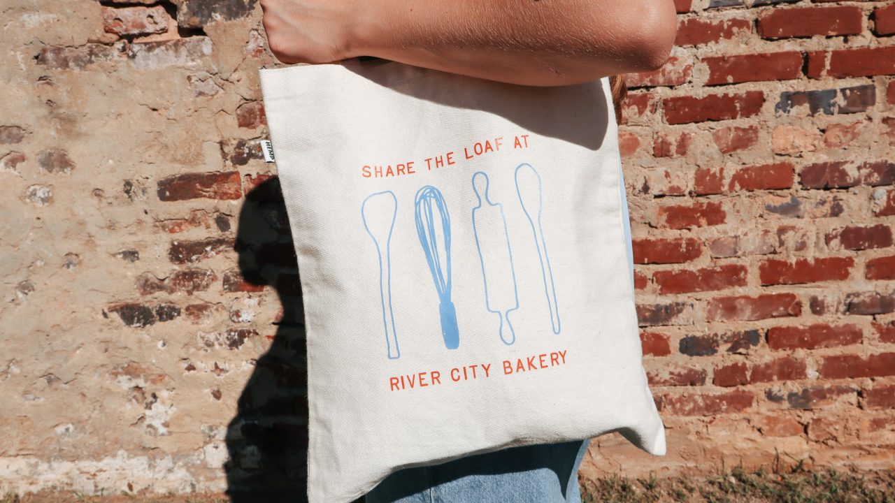 A person holds a fabric tote bag featuring blue illustrations of baking tools and the text Share the Loaf at River City Bakery in red letters.