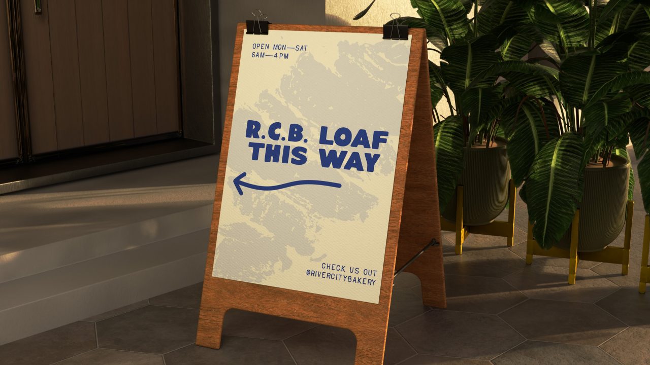 A wooden sandwich board sign reads R.C.B. Loaf This Way with a left arrow. It lists bakery hours and social media handle @rivercitybakery.