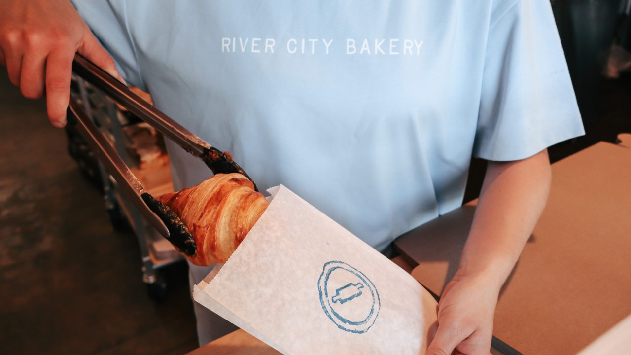 A person in a River City Bakery shirt uses tongs to place a croissant into a branded paper bag.